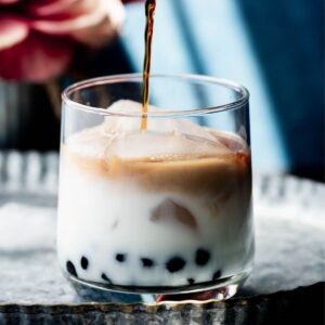 hero image of caramel milk boba tea - tea is going into a clear class with milk and boba. The glass is on a silver plate.