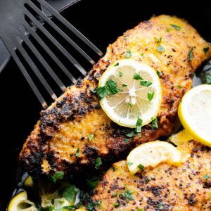 Photo of lemon pepper chicken with a lemon and parsley in a black pan.