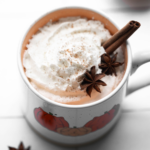 The top of a mug with pumpkins on it, filled with chai latte, whipped cream, and garnished with a cinnamon stick.
