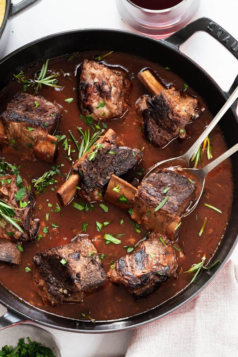 Short ribs in a braiser with fresh herbs and spoons. 