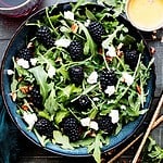Blackberry salad with arugula in a blue bowl with homemade dressing on the side.